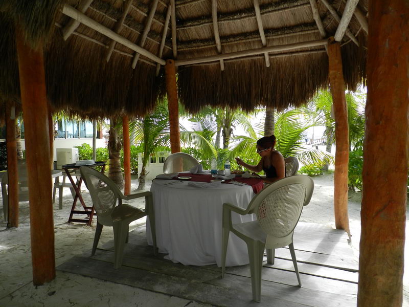 A woman sits alone at a round table set for dining under a large thatched palapa on a beach. White plastic chairs surround the white-clothed table; palm fronds and sandy ground visible beyond the shaded structure.