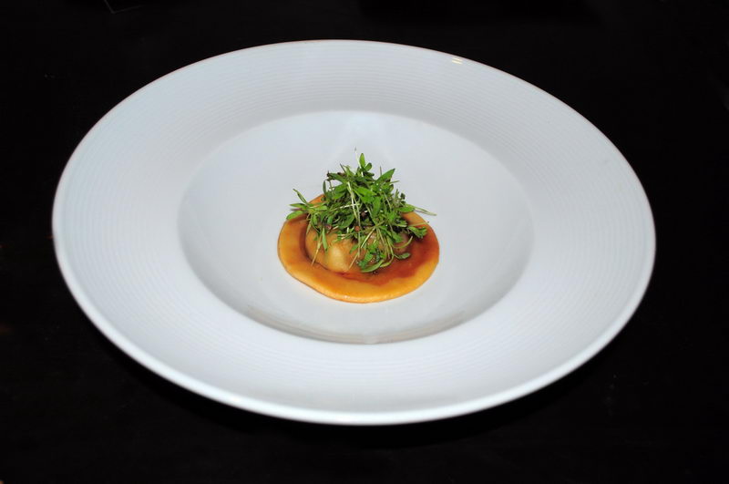Minimalist fine-dining dish in a deep white bowl: a single round ravioli in light brown sauce topped with a tall tuft of green micro-greens, set against a black background. Elegant restaurant plating.