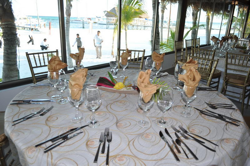 Elegant round banquet table set with white patterned cloth, gold napkins folded into rosettes, wine glasses, and cutlery, beside floor-to-ceiling windows overlooking a beach and marina. Gold Chiavari chairs surround the table.