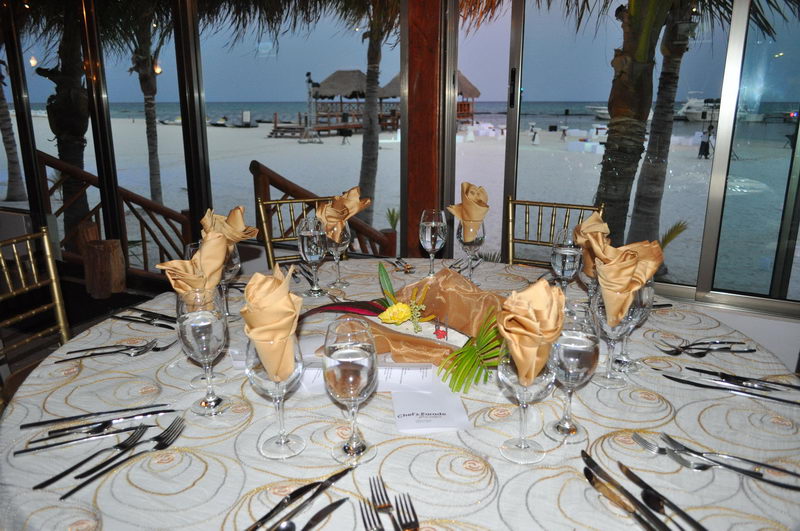 Round dining table with white swirl-patterned cloth and gold rose-folded napkins, wine glasses and cutlery, set by large windows at dusk overlooking a beach with docked yachts. Gold Chiavari chairs and warm interior lighting.