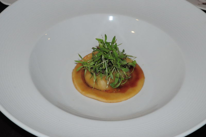 Fine-dining appetizer served in a deep white bowl: a round pastry or dumpling in brown sauce crowned with a tall tuft of green herbs and micro-greens. Photographed in dim restaurant lighting.