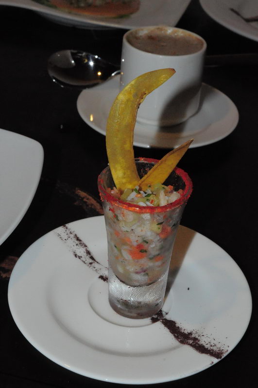 Seafood ceviche or salad served in a tall shot glass with a red-rimmed edge, garnished with two curved fried plantain or tortilla chips. Set on a white plate dusted with spice; a coffee cup sits behind on a dark table.