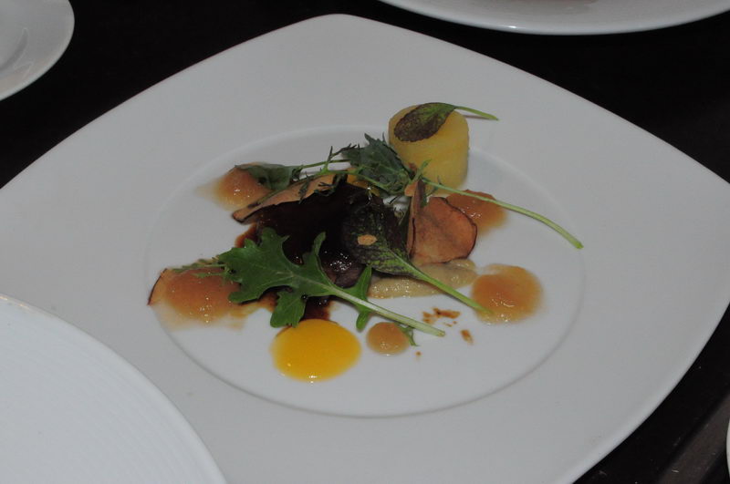 Artistic fine-dining plate on white square dish: a small yellow potato cylinder, arugula leaves, thin dark chips, and dots of orange and brown sauces artfully scattered. Dark background, dim restaurant lighting.