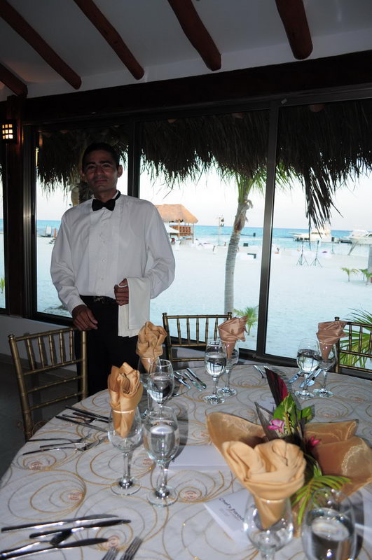A waiter in a white shirt and black bow tie stands beside an elegantly set round table with gold napkins and glasses, inside a beachfront restaurant with windows overlooking a turquoise marina and palm trees at dusk.