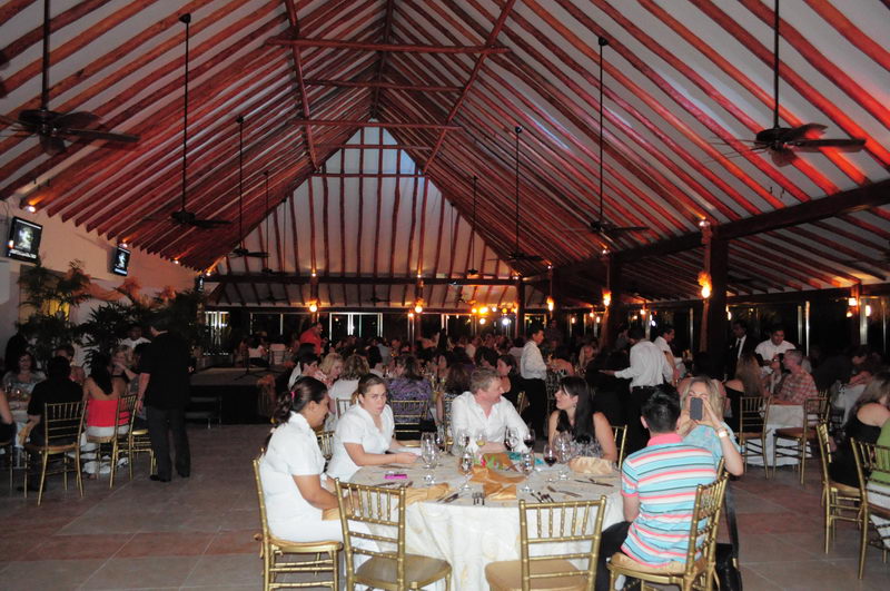 Crowded evening banquet inside an open-sided hall with a peaked wood-beamed roof lit in warm tones. Many guests sit at cloth-covered tables with wine glasses; staff in white move among them. Lively dinner-event atmosphere.