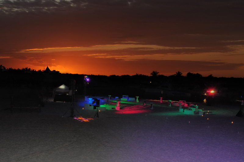 Nighttime beach event setup photographed at sunset: an orange-glowing horizon over a dark tree line, with a lit lounge area of glowing furniture, colored stage lights, and a canopy tent on the sand. Dim, atmospheric.