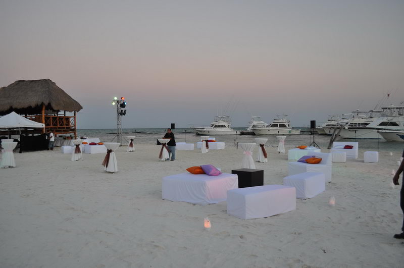Beach event at dusk with white cocktail sofas, cloth-draped high tables, and colored pillows arranged on the sand. Stage lighting rigs stand ready; sport-fishing yachts are docked along the shore under a pale pink sky.