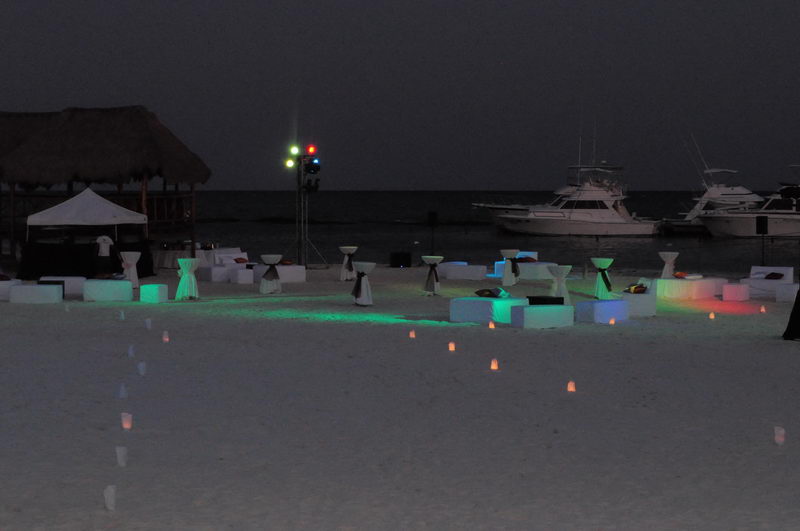 Nighttime beach event with glowing illuminated cube furniture, cloth-draped cocktail tables, and candle luminaries lining a path on the sand. Colored stage lights and docked sport-fishing yachts sit in the dark background.