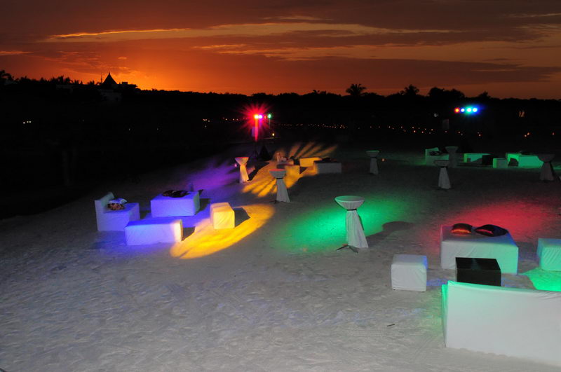 Beach party setup at sunset with glowing white lounge cubes and cocktail tables scattered on the sand, colored stage lights casting green, yellow and purple beams. An orange sky glows over a dark palm tree line.