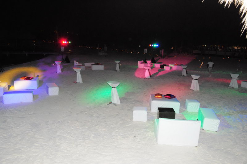 Nighttime beach event with illuminated white lounge cubes, draped cocktail tables and colored pillows spread across the sand. Multicolored stage lights and distant shore lights glow under a black sky.