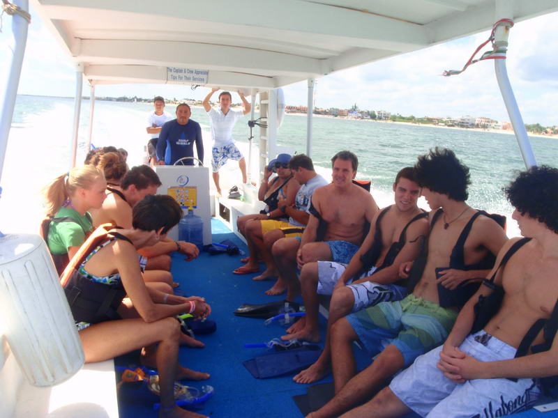 Group of tourists in life vests seated aboard a covered blue-decked tour boat underway, holding snorkel gear and fins. Two crew members stand at the center; a beach town shoreline passes behind under a bright sky.