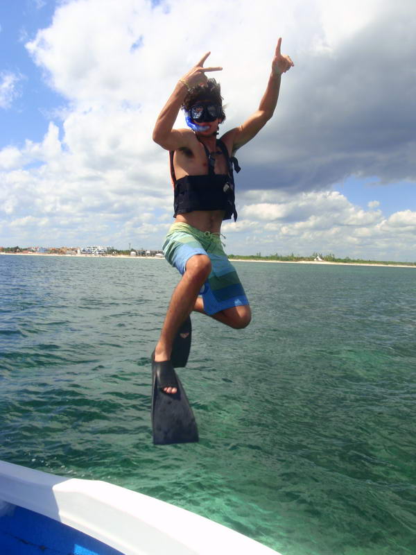 A young man in board shorts, snorkel mask and life vest leaps off a boat's bow into the sea, arms raised making hand signs, fins pointed down. Turquoise water, a distant beach shoreline and cloudy blue sky behind.