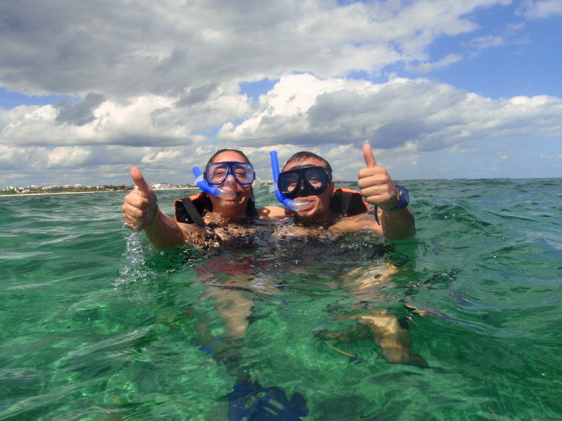 A smiling couple in snorkel masks float at the surface of clear green-blue water, both giving thumbs-up to the camera. A beach shoreline and cloudy blue sky are visible in the background.