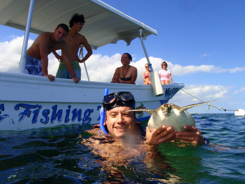 A snorkeler in the water holds up a small sea turtle toward the camera beside a white tour boat labeled 'Fishing'. Several young people watch from the boat's deck under a bright blue sky with clouds.