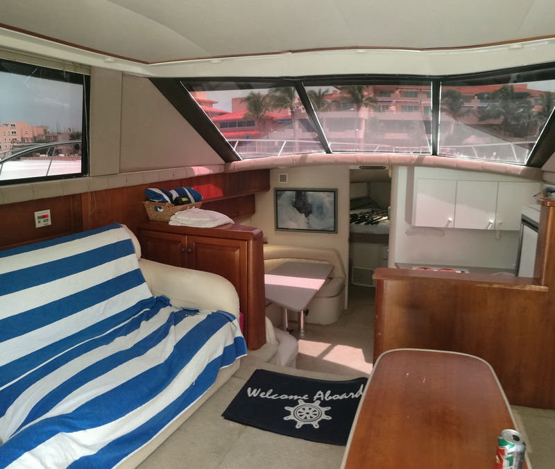 Interior salon of a flybridge motor yacht with a cream leather settee draped with a blue-and-white striped towel, wood cabinetry, a 'Welcome Aboard' mat, a nautical framed print and large wraparound windows showing a resort outside.