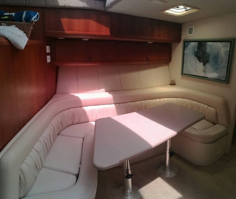 Interior dinette of a motor yacht with a cream leather U-shaped settee around a white table, wood paneling, an overhead cabinet, a framed lighthouse print and towels in a basket. Sunlight streams across the cushions.