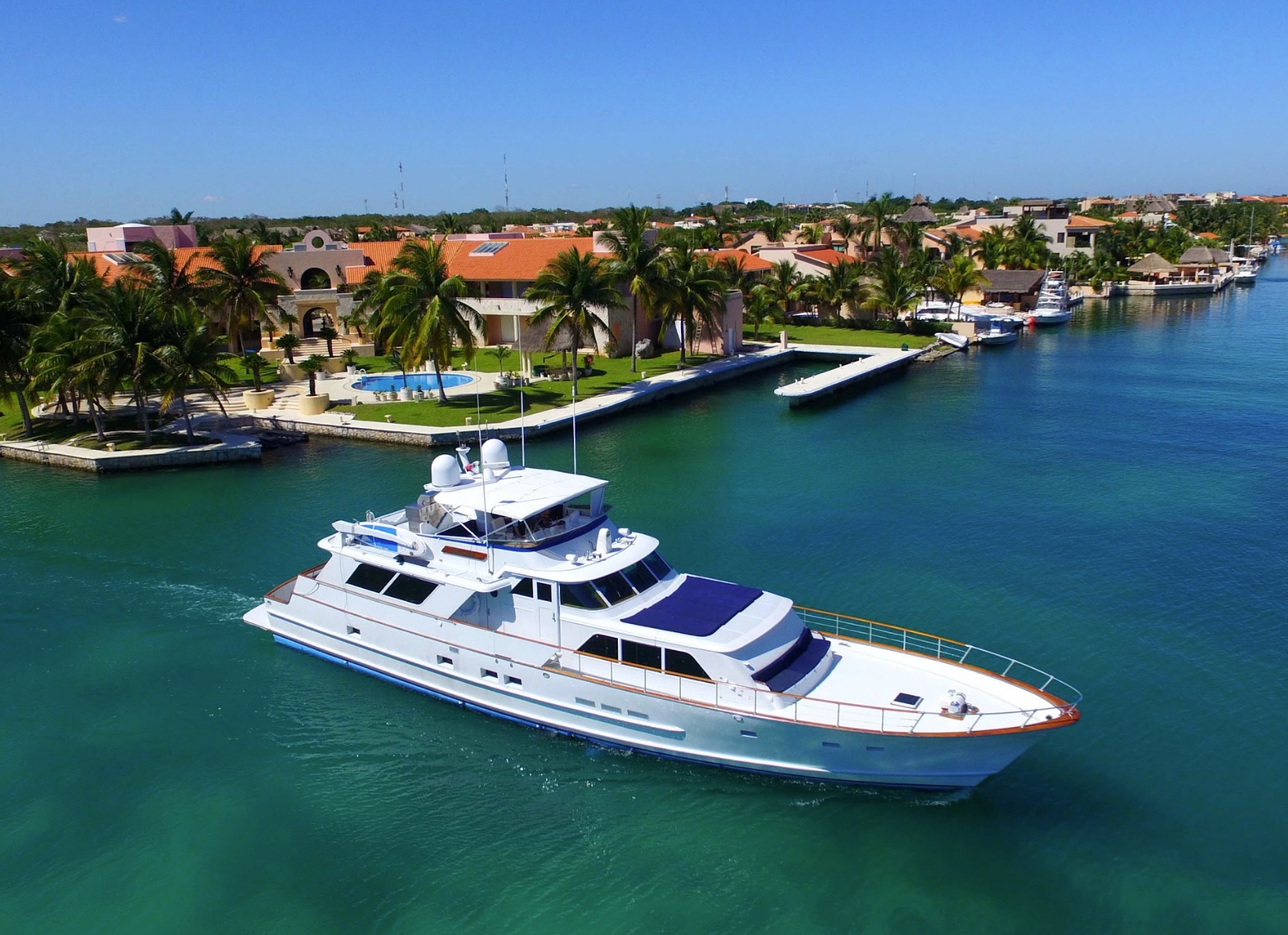 Dramatic low-angle GoPro view of a gray-hulled explorer motor yacht's bow rising from turquoise water. The flared hull, wood cap rail and portholes dominate the frame against a blue sky with puffy clouds.