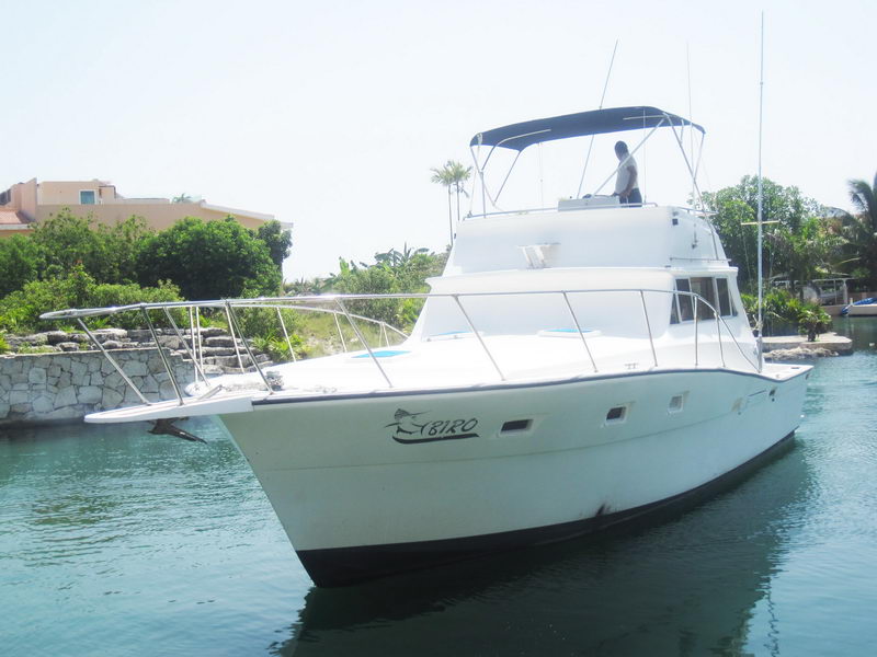 White flybridge sportfish yacht 'Biro' viewed bow-on beside a rock wall, a crew member standing on the flybridge. Bimini top, bow rails and hull-side lettering visible; marina condos and palms behind under a clear sky.