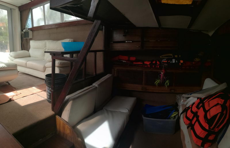 Dim interior cabin of an older sportfishing boat with white leather sofas, a wood ladder to the flybridge, and orange life jackets stowed against a wood bulkhead. Cluttered utilitarian space with a blue basin and storage bins.