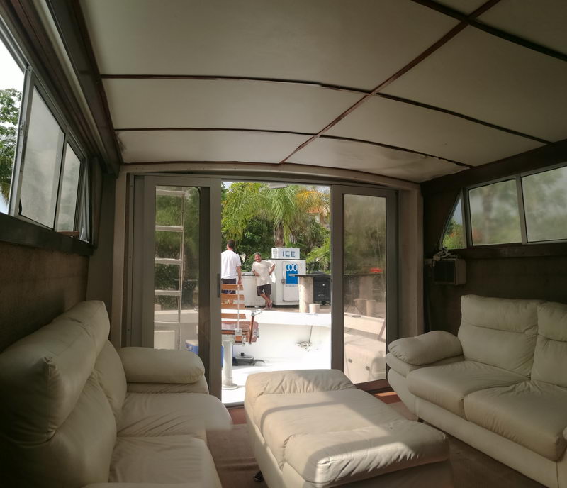 Interior salon of a sportfishing boat with two white leather sofas and an ottoman, looking through open sliding doors to the aft cockpit where two men stand near an ICE machine dockside. Padded headliner and side windows frame the space.