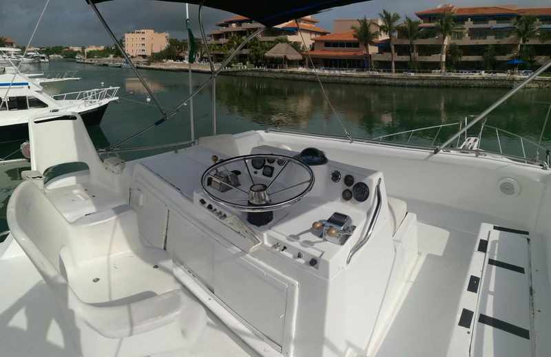 Flybridge helm of a sportfishing yacht with a black steering wheel, gauges and a white console, under a bimini top. Overlooks calm green marina water with docked sportfish boats and waterfront condos beneath a cloudy sky.