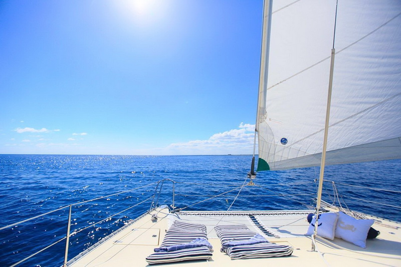 Foredeck view from aboard a sailing catamaran looking forward past white striped cushions and pillows on the deck, the large white sail to the right and the sun high over deep blue open ocean stretching to the horizon.