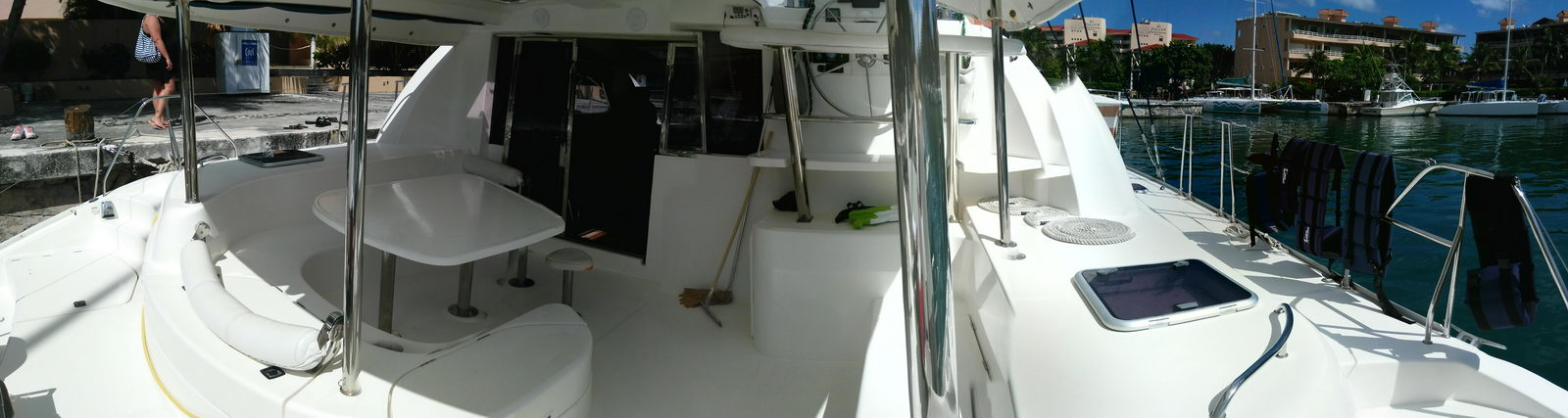 Panoramic aft cockpit of a Leopard catamaran under its hard top: white molded dinette table and bench seating, deck brooms, dive fins, hatches into the hulls, docked beside a stone quay with sailboats and clear water behind. Sunny.