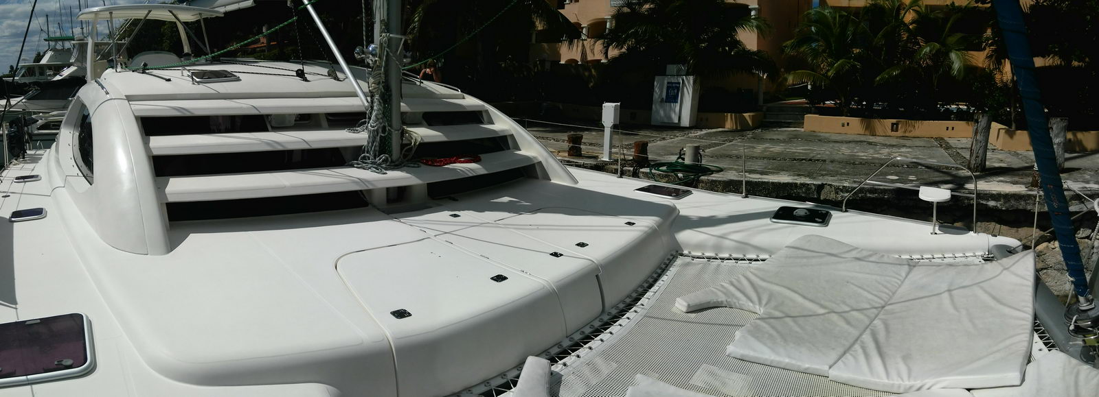 Panoramic foredeck of a Leopard catamaran showing the raised coachroof with stepped windows, white deck between the hulls, the trampoline netting with cushions laid out, and the mast base. Docked by a quay with palms and villas, bright sun.