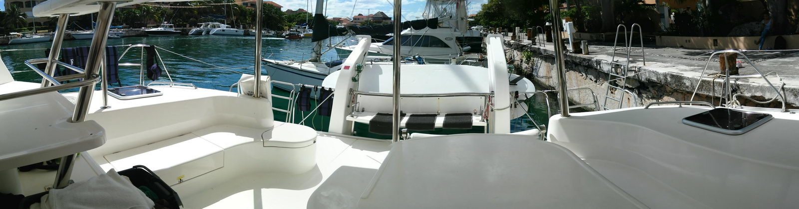 Panoramic view across the foredeck of a Leopard catamaran between the twin hulls, with white deck seating, hatches and a boarding ladder, docked stern-to in a marina among other catamarans and motor yachts. Clear water, sunny day.