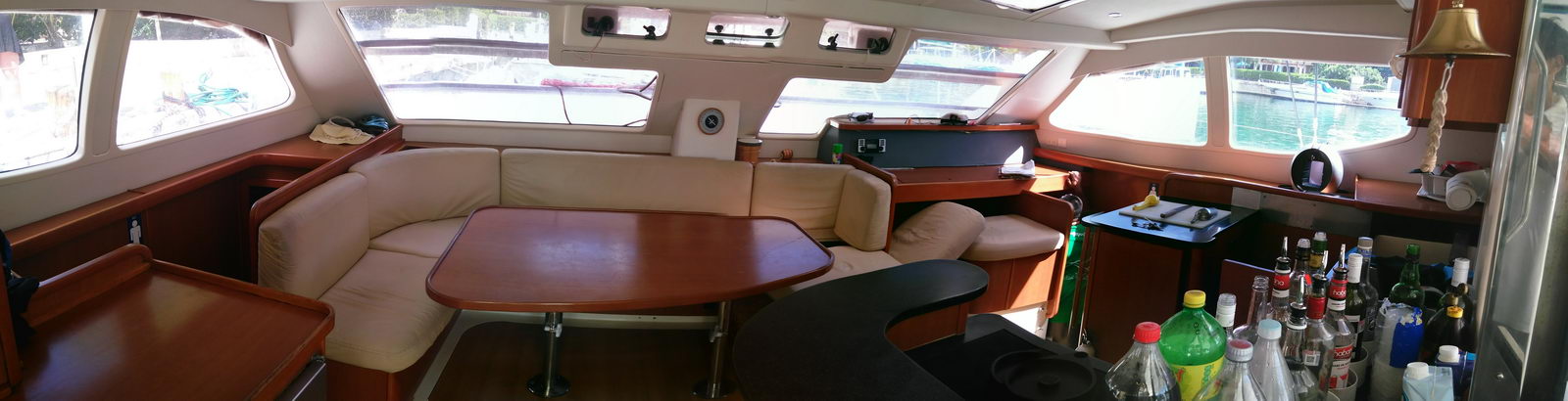 Panoramic interior salon of a sailing catamaran with cream leather U-shaped settee, a varnished wood dining table, wraparound windows showing a marina outside, and a bar counter cluttered with liquor and soda bottles.