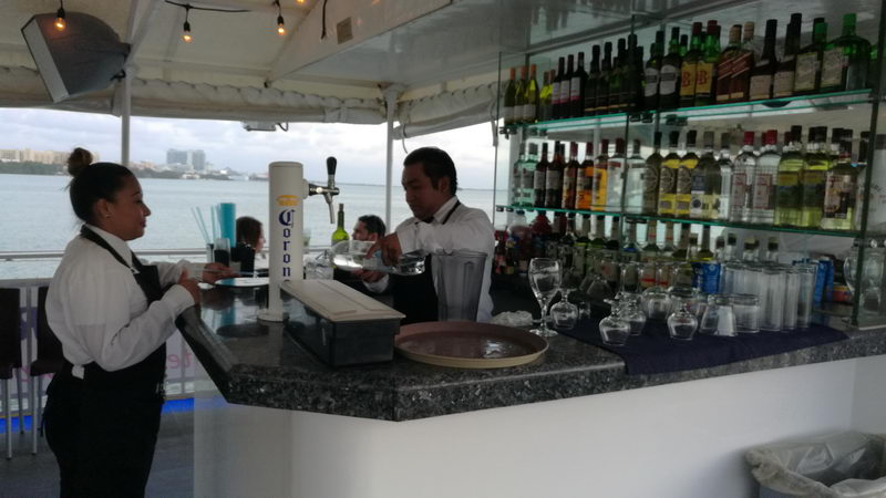 Onboard bar of the dinner-cruise boat with a bartender preparing drinks and a waitress at the counter, shelves of liquor bottles, Corona beer tap, glassware, and the Cancun hotel-zone skyline across the water at dusk.