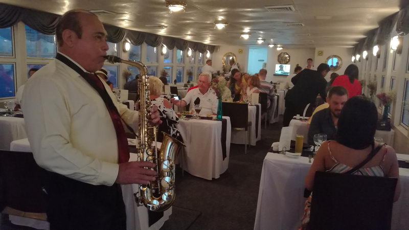 Saxophonist in white shirt and vest playing live music between tables in the boat's window-lined dining room while dinner guests eat at tables with white tablecloths and dark runners.
