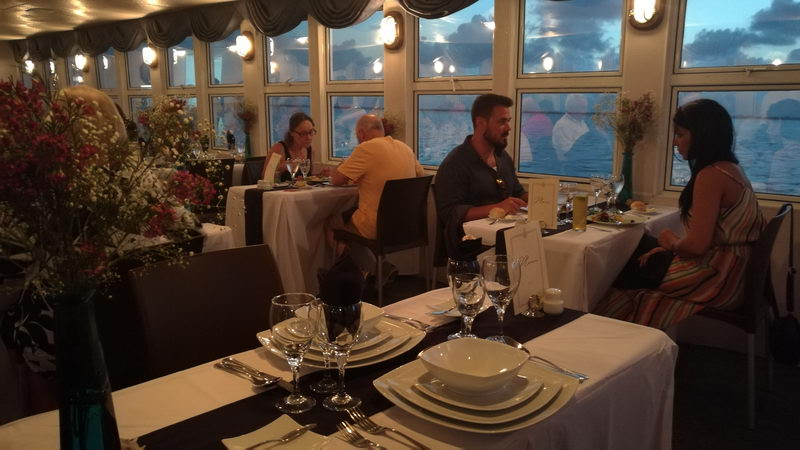 Guests dining at set tables in the paddle boat's dining room, panoramic windows showing dusk light on the lagoon, white tablecloths with dark runners, square white china and fresh flower arrangements.