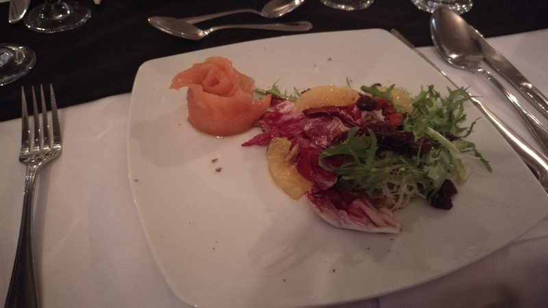 Dimly lit appetizer plate served on white square china: smoked salmon rolled into a rose shape with mixed greens, radicchio, citrus segments and dried fruit, silverware and wine glasses around it.
