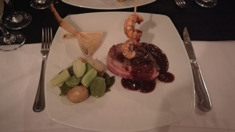 Main course on white square plate in dim dinner lighting: bacon-wrapped filet mignon in dark sauce topped with a shrimp skewer, steamed vegetables and baby potatoes, with a corn-husk garnish.