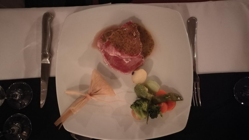 Plated meat course in peppercorn-style sauce with broccoli, carrots and asparagus and a decorative corn-husk bow, photographed from above on a white table with knife and fork, dim evening light.