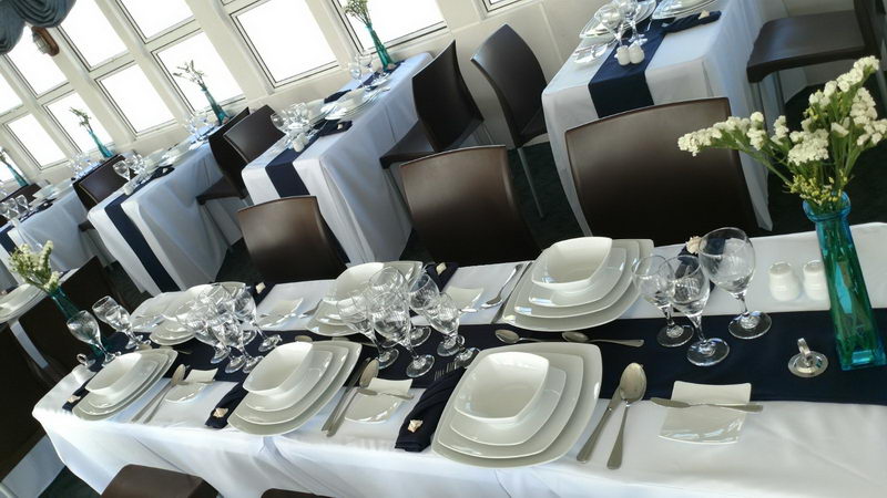Dining room of the cruise boat set for service in daylight: rows of tables with white tablecloths, navy runners, square white china, polished glassware, brown chairs and turquoise vases with white flowers by the windows.