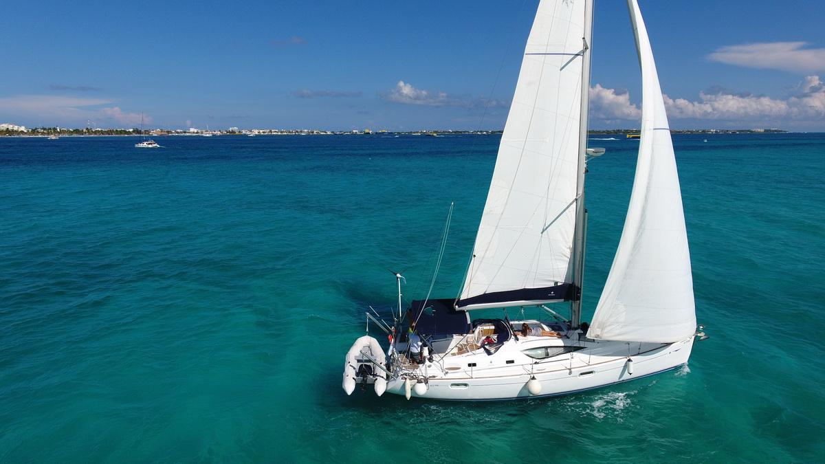 White Jeanneau sailboat under full main and genoa gliding on turquoise-blue sea, dinghy stowed at the bow rail, crew visible in the cockpit, low hotel-lined coastline on the horizon under a blue sky.