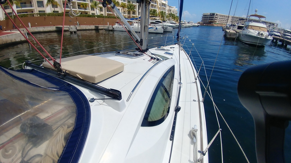 View from a sailboat's sprayhood across the coachroof with a beige sun cushion, leaving a marina channel lined with moored motor yachts, catamarans and waterfront condo buildings on a sunny day.