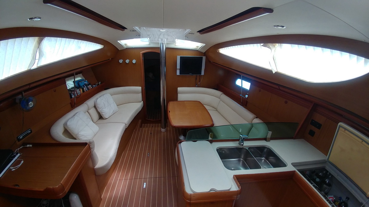 Wide-angle salon interior of a Jeanneau sailboat: white leather settees port and starboard, cherry woodwork, wood dining table, flat-screen TV on the bulkhead, double stainless galley sink and open fridge in the foreground.