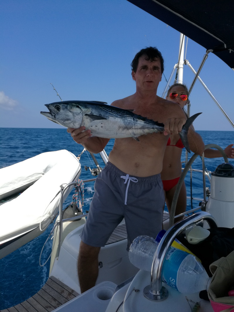 Shirtless man in gray swim shorts on a sailboat's stern holding a skipjack tuna with both hands, a woman in a red bikini standing at the helm behind him, dinghy stowed on deck, calm blue sea.