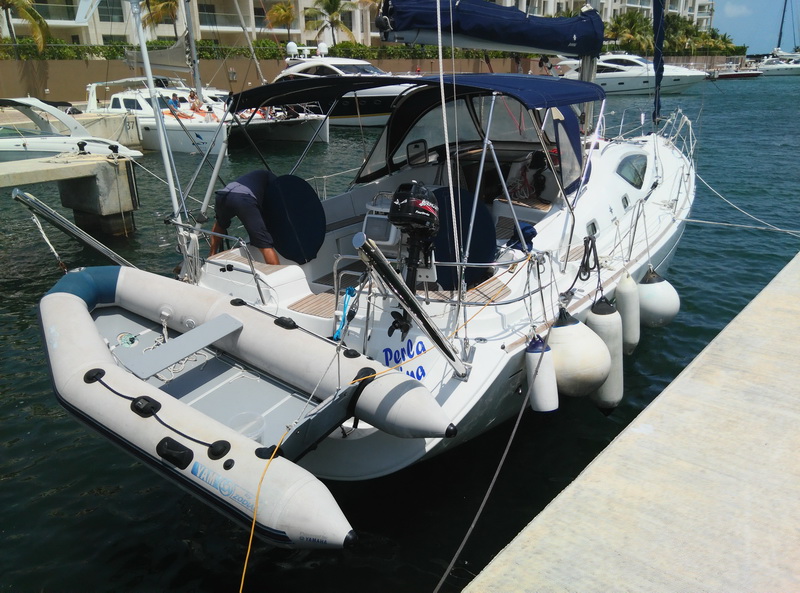 Stern view of a white sailboat docked at a marina, gray inflatable dinghy with outboard tied behind, crew member working on deck, white fenders along the hull and hotels with palm trees behind.