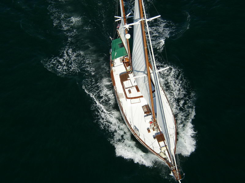 Top-down aerial of a classic white-hulled ketch under sail leaving a wake in dark water, teak deck, skylights and green deckhouse awning seen from directly above the masts.