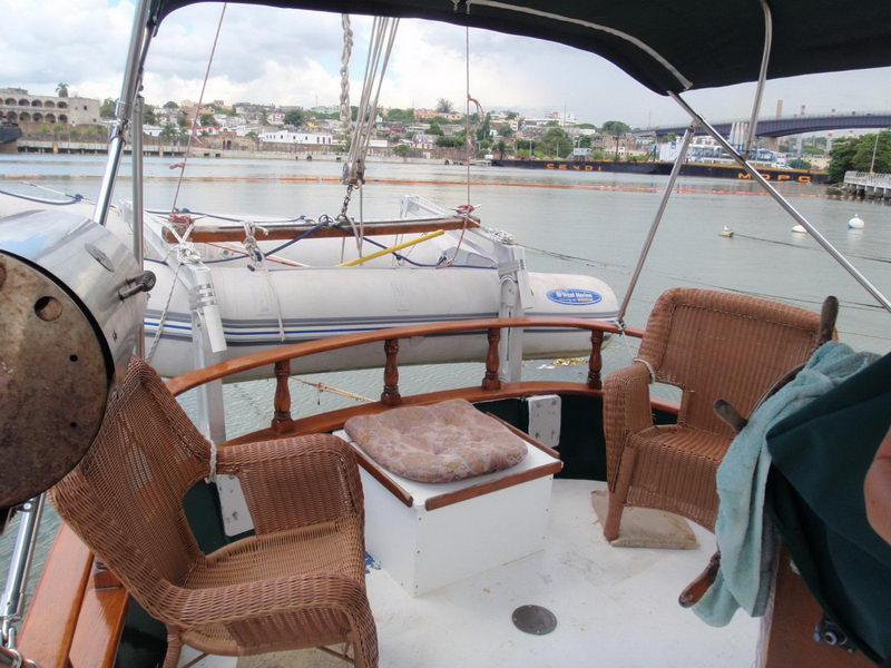 Aft deck of a classic sailboat with two wicker armchairs and a cushioned box seat, varnished wood rail, gray inflatable dinghy stowed on davits, riverside town and bridge in the background on an overcast day.