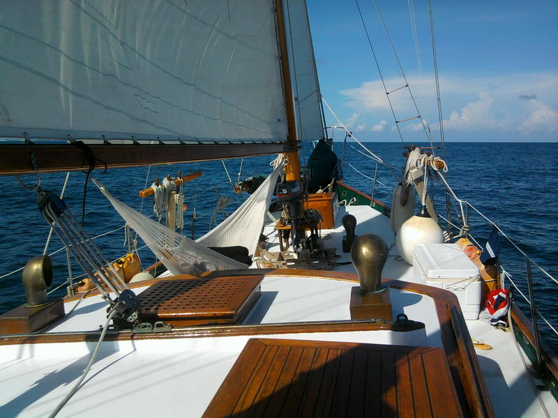 Looking aft along the white foredeck of a classic ketch under sail, varnished teak hatches and skylight, a white mesh hammock strung by the mast, brass vents and an outboard motor mounted midship, deep blue sea beyond.