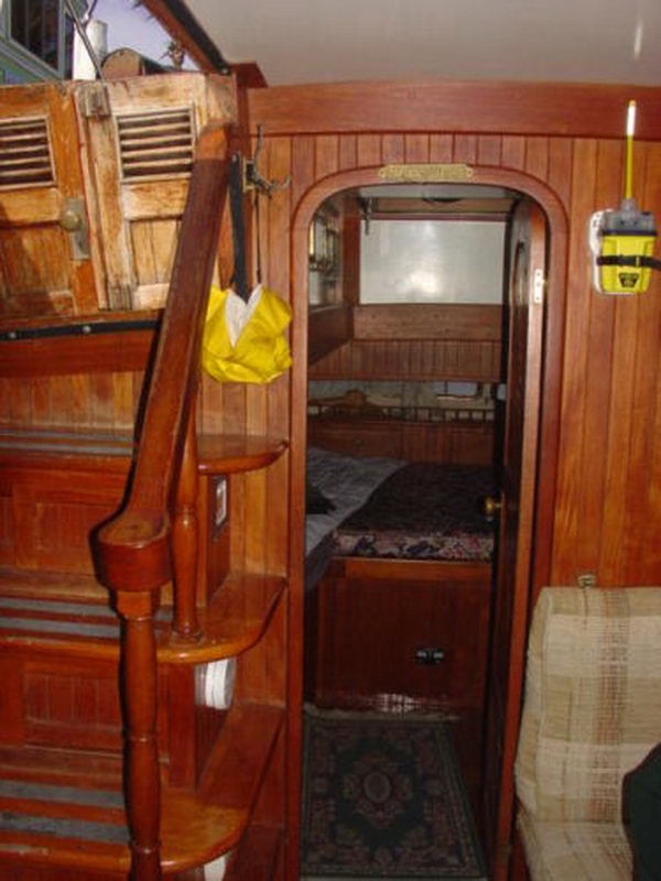 Classic sailboat interior in dark varnished mahogany: companionway steps with louvered doors on the left and an arched doorway leading to a sleeping cabin with a double berth and oriental rug.