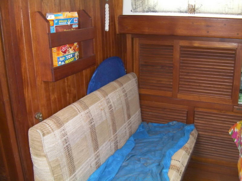 Corner of a wood-paneled sailboat salon with a plaid upholstered settee partly covered by a blue tarp, wooden magazine rack holding cereal boxes and louvered locker doors behind.