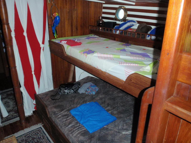 Sailboat sleeping cabin with a wooden-framed upper bunk with colorful bedding and a wide lower berth below, red-and-white striped curtain, toy parrot hanging by the bunk and a brass porthole on the striped wall.