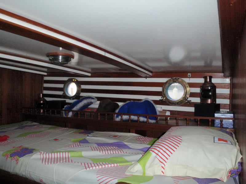 Wide double berth in a classic sailboat cabin with colorful patterned bedding, red-and-white striped bulkhead with two brass portholes, wooden rail shelf with a copper jug and ship lantern.
