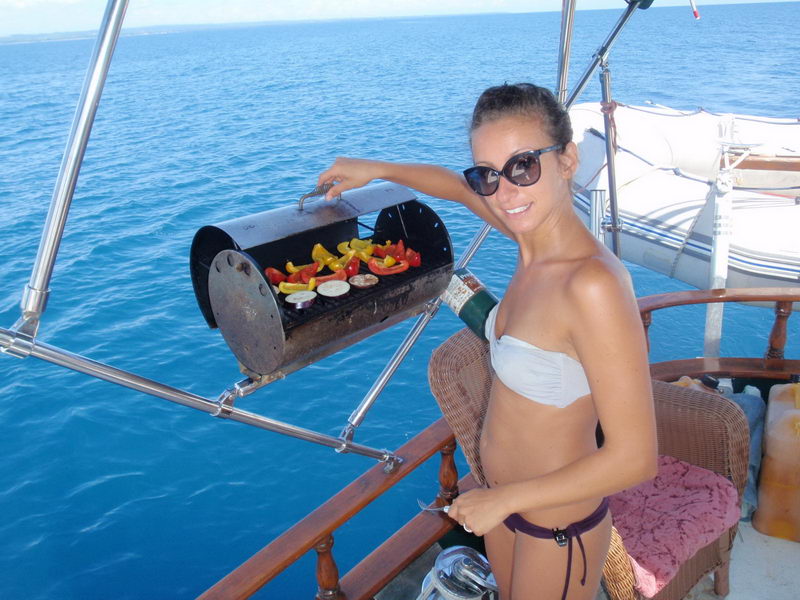 Smiling woman in a white bikini top and sunglasses cooking peppers and vegetables on a rail-mounted drum barbecue at the stern of a boat, deep blue sea and an inflatable dinghy behind her.